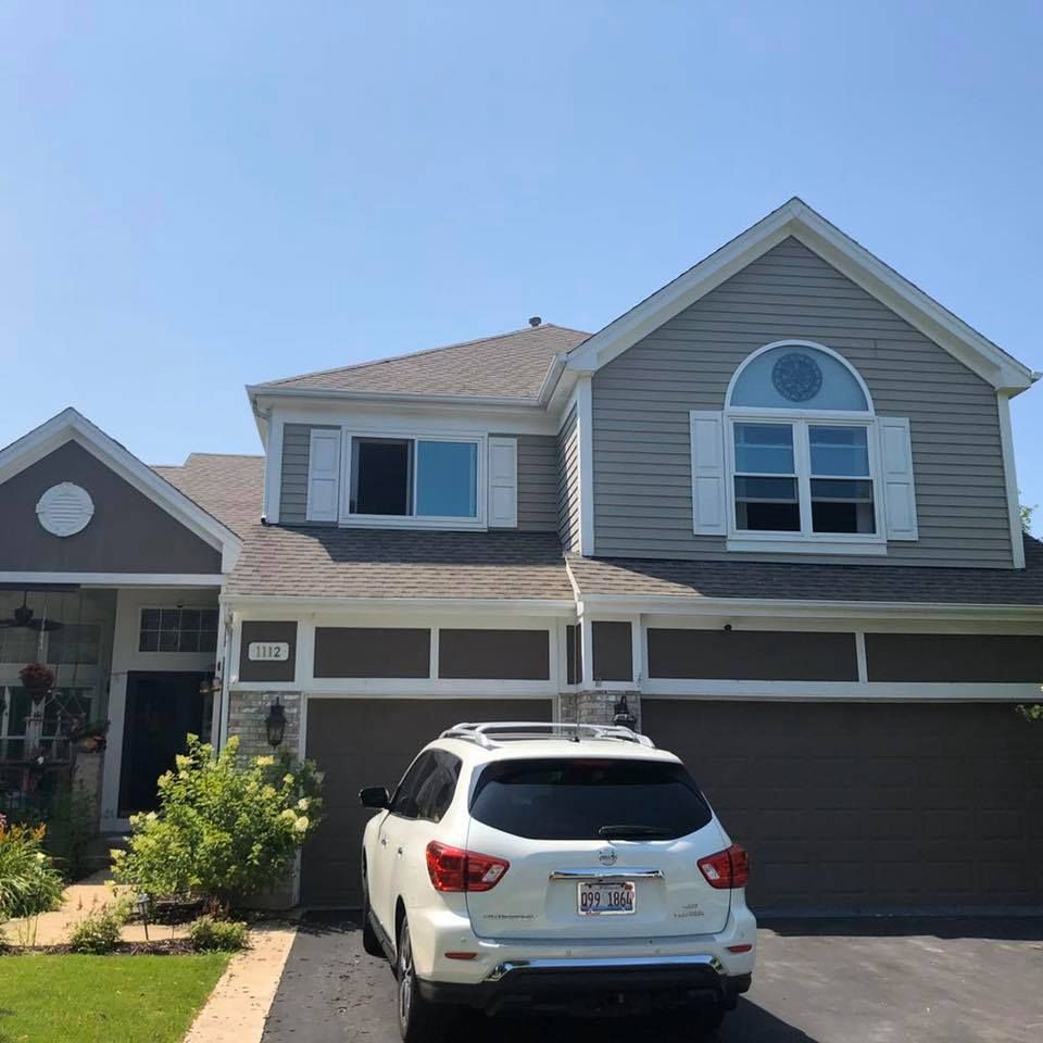 A two-story gray and brown house with a white SUV parked in the driveway on a sunny day.