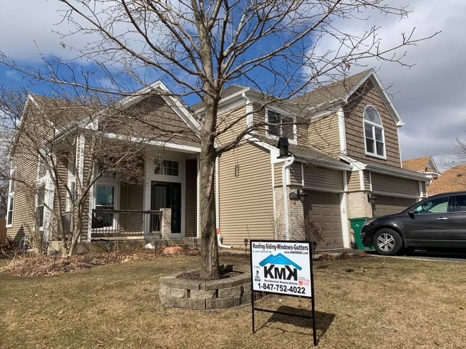 A tan two-story house with a lawn, a central tree, a driveway with a dark SUV, and a business sign in the foreground.