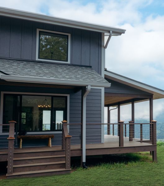 A modern house with dark gray vertical and horizontal siding and a wide wooden deck overlooking a distant mountain view.