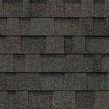 A close-up of dark grey, overlapping architectural asphalt roof shingles with a textured, layered appearance.