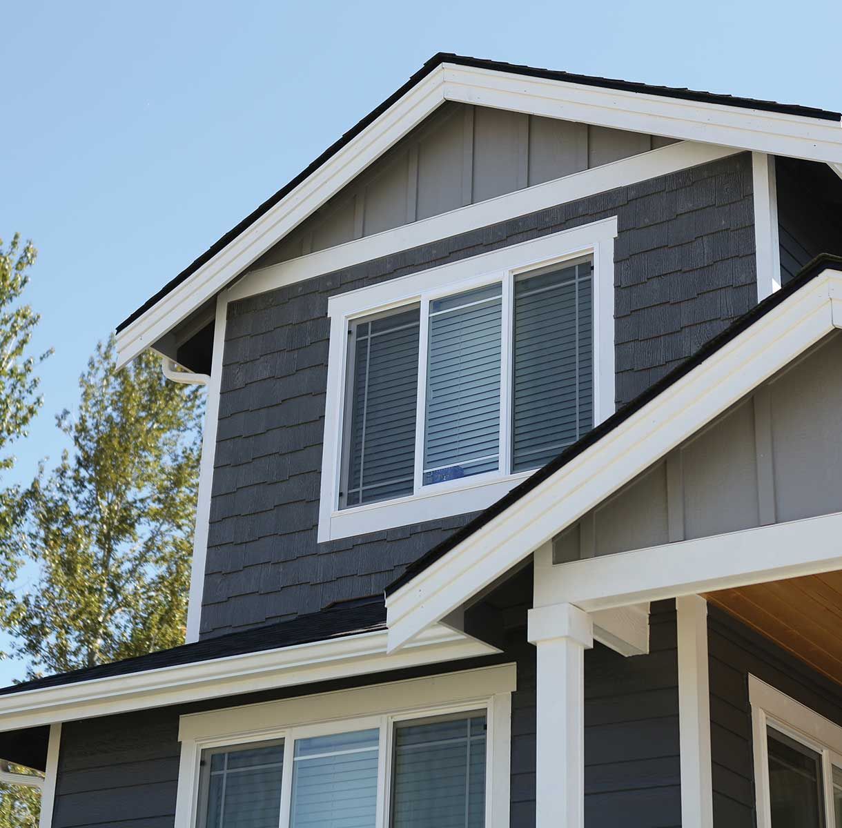 A two-story house exterior with dark gray shingles, white trim, and white-framed windows under a clear blue sky.