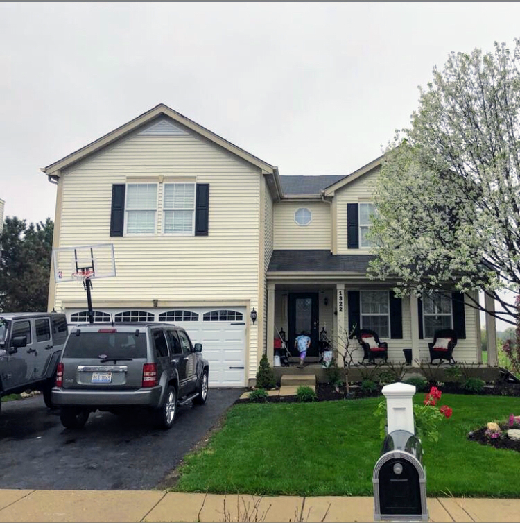 A two-story beige house with black shutters, a two-car garage, a driveway with two SUVs, and a blooming tree in the yard.