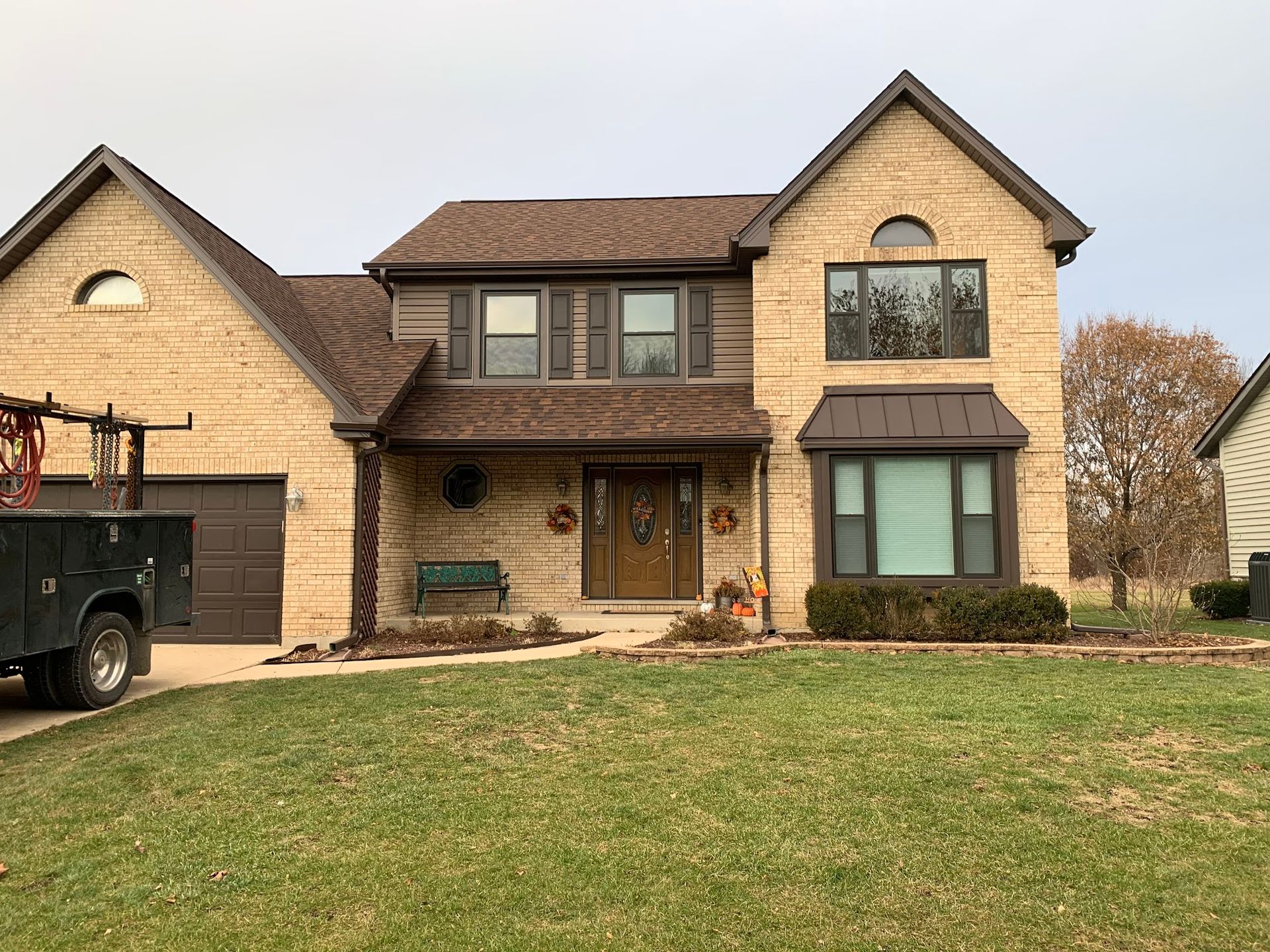 Two-story, tan brick house with a dark brown roof and trim, a front porch, and a driveway with a work truck parked on side.