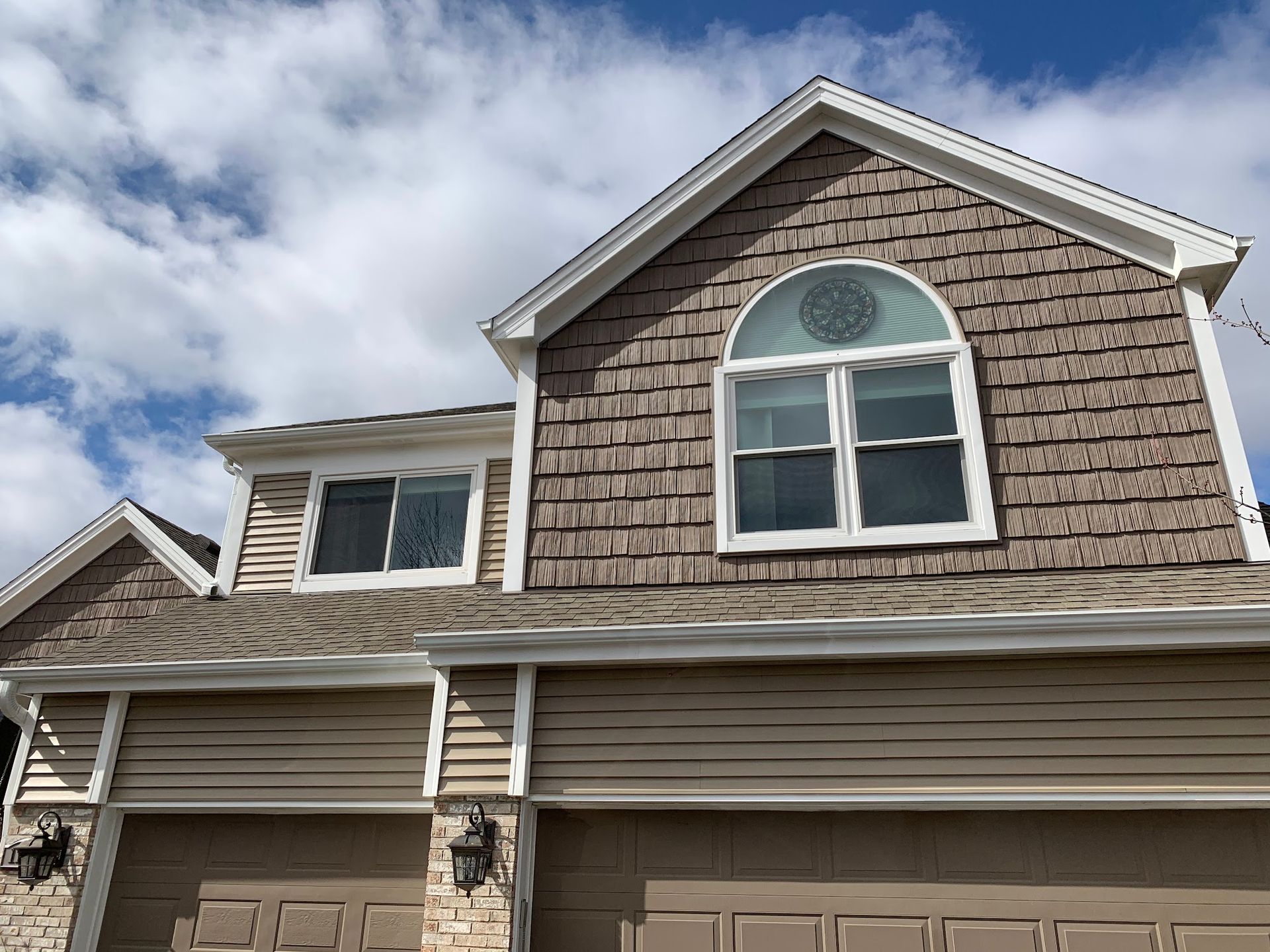 A two-story suburban house exterior with tan horizontal siding, brown shake shingles on the upper gable, and garage doors.