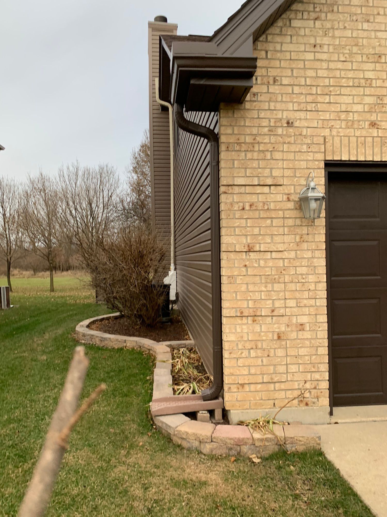 Exterior view of a tan brick house corner with a brown downspout and a stone-walled garden bed next to a lawn.