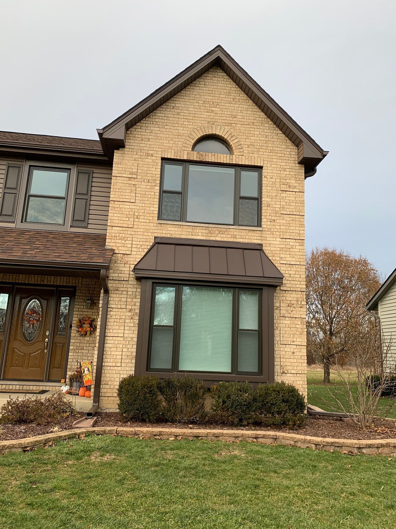 Two-story home exterior with tan stone facade, dark metal roof trim, a prominent bay window, and a brown front door.
