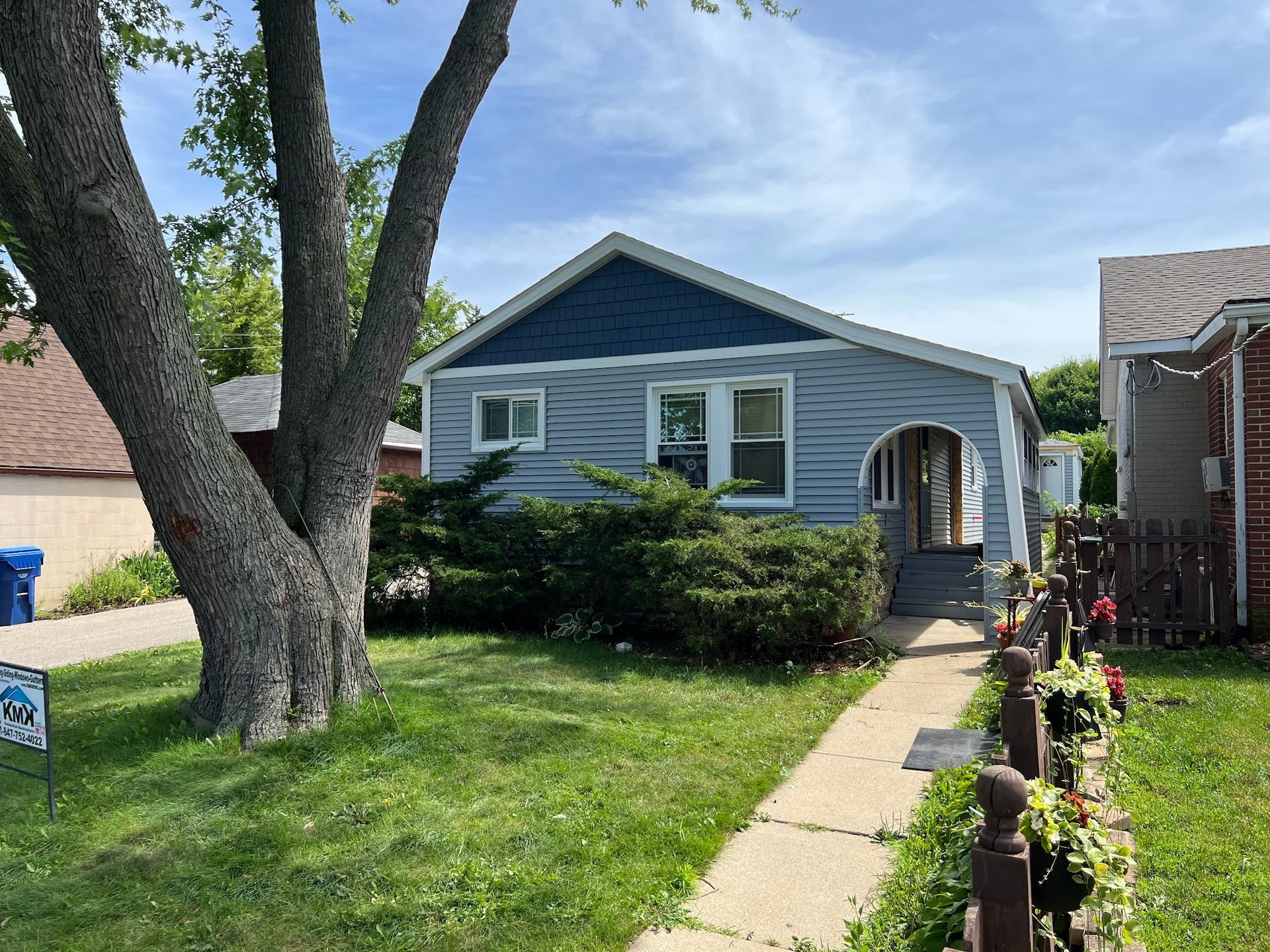 A light blue, single-story house with a white arched entryway and a large tree in the front yard.