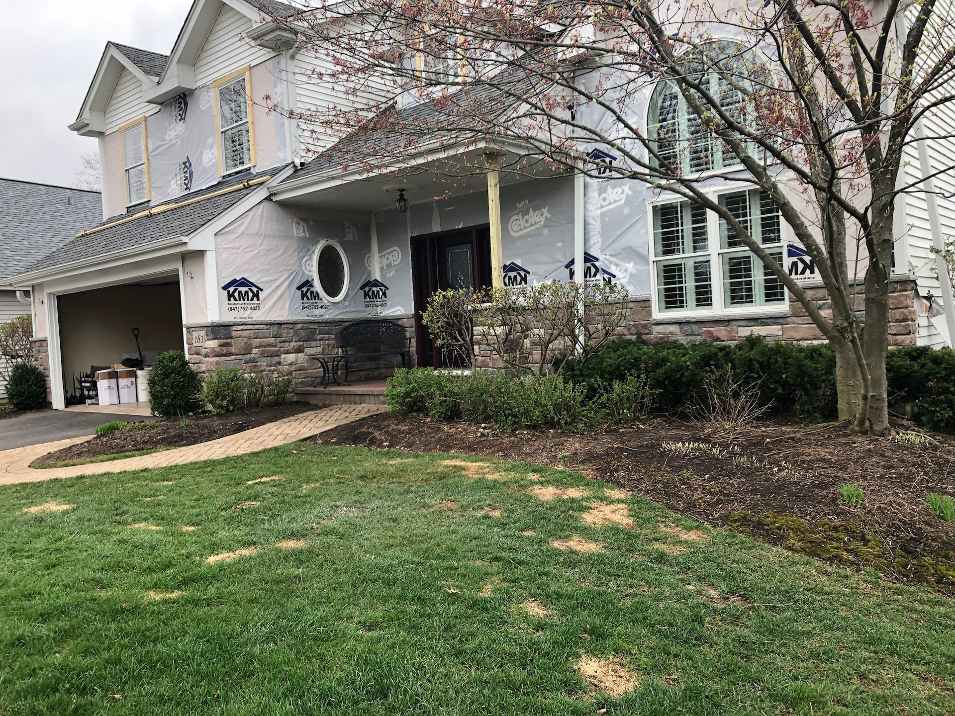 A two-story house undergoing renovation with visible weather-resistant barriers on the siding and a stone-veneer base.