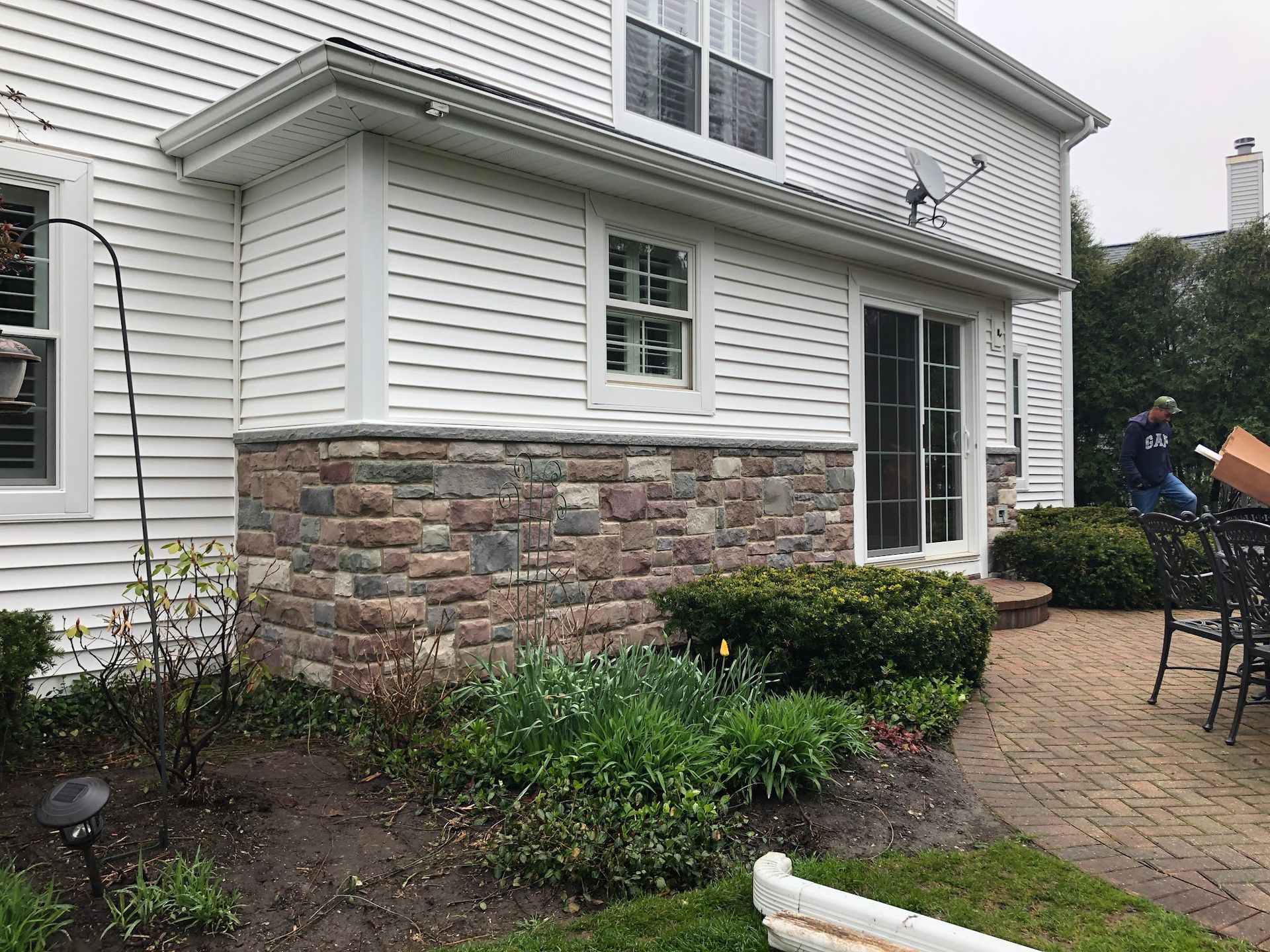 Side view of a white house with stone siding on the lower level, a patio, landscaping, and a person in the background.