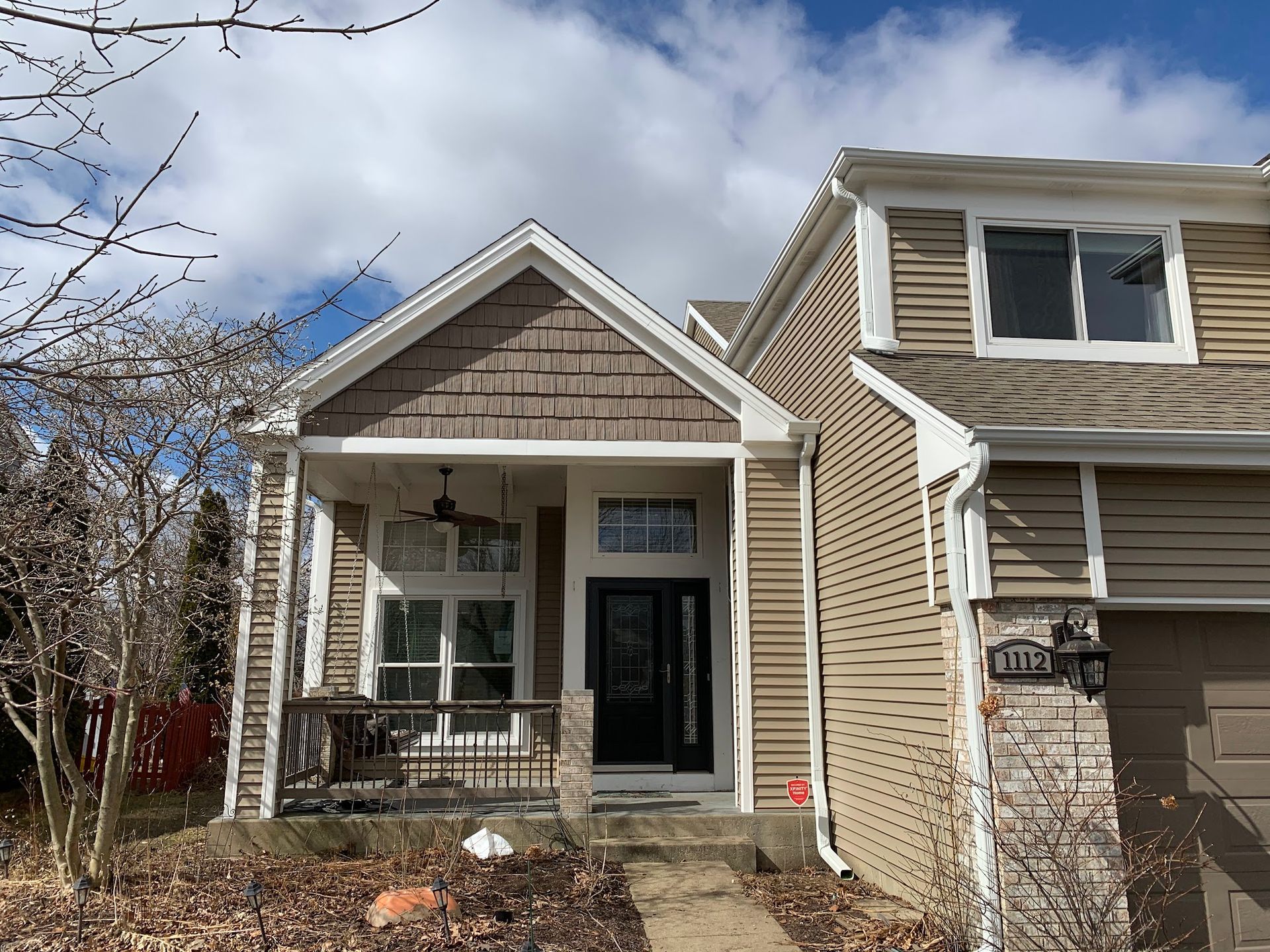 A tan house with a front porch, shake-style siding on the gable, a dark door, and an attached garage under a blue sky.