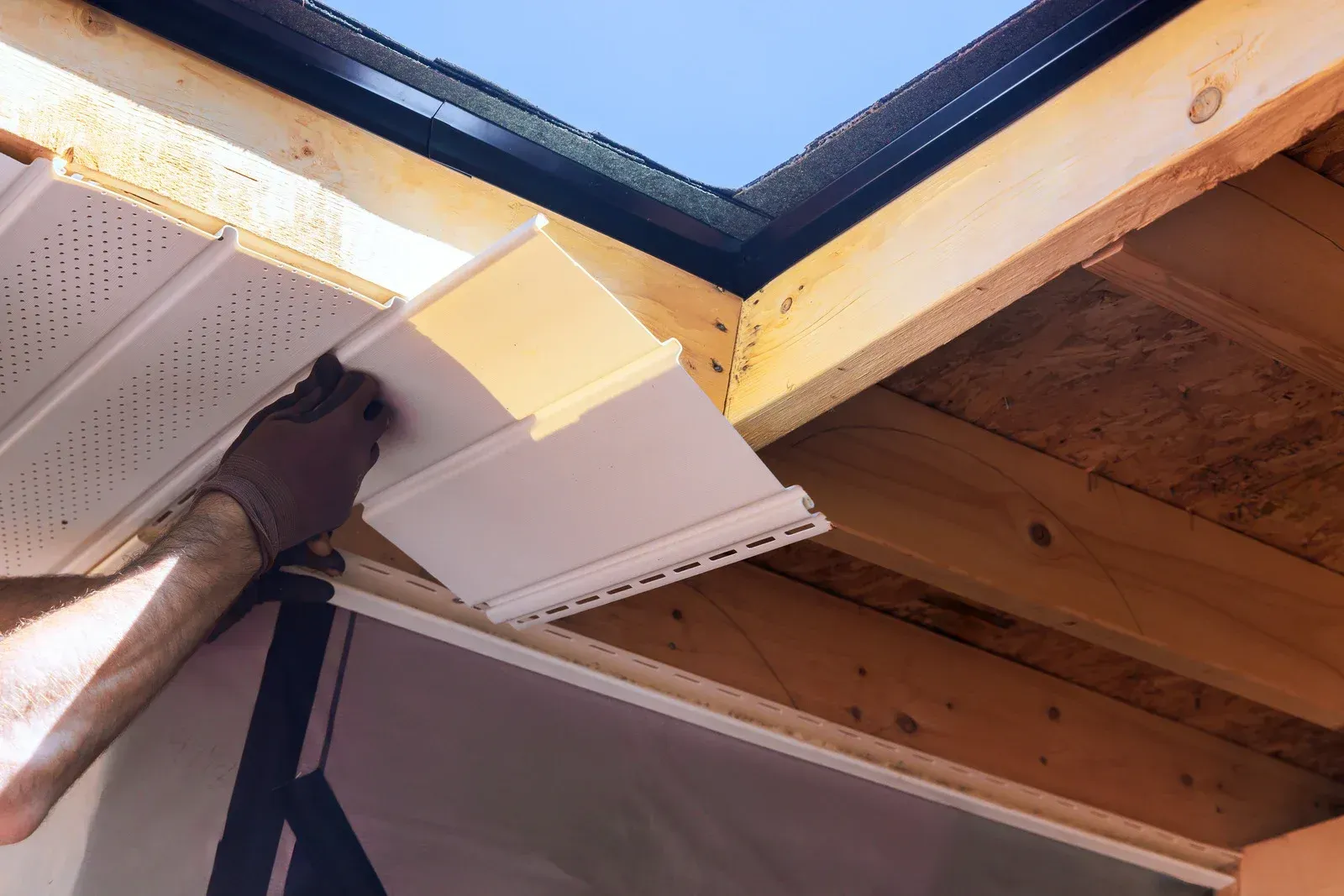 Side view of a light-colored house with dark brown trim, a red roof, a white gutter, and a wooden privacy fence.