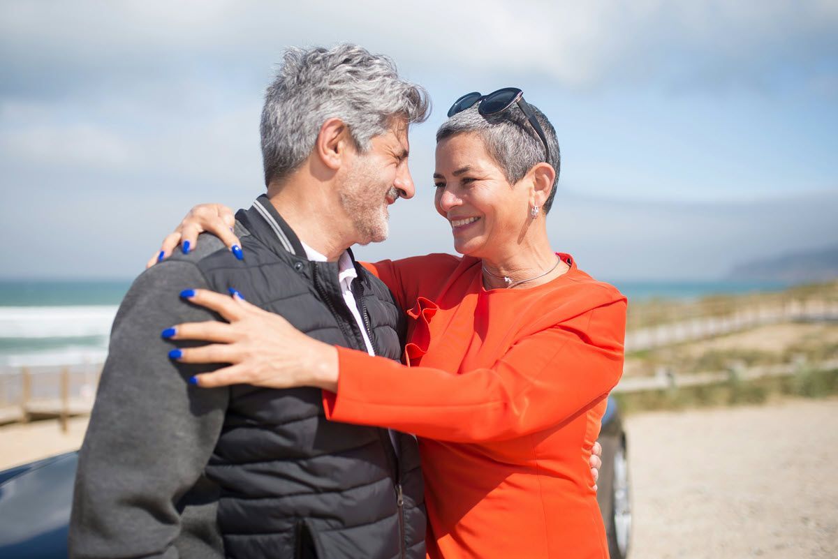 A retired couple smile and embrace outdoors near the beach.