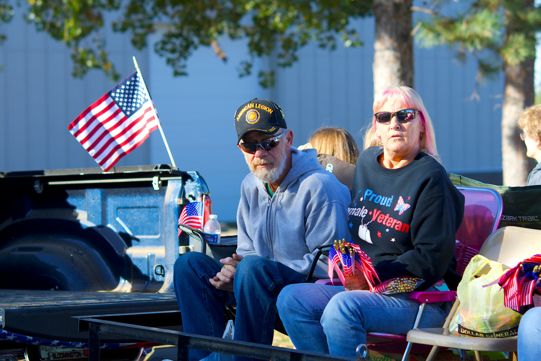 Two people seated outdoors beside an American flag, with a car and bags nearby.