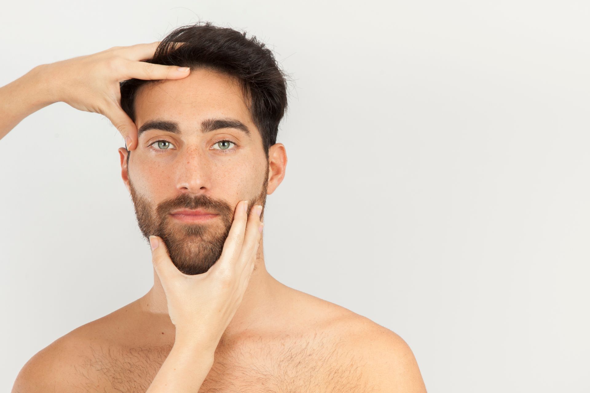 Hands gently frame the face of a man with dark hair and a beard against a plain white background.
