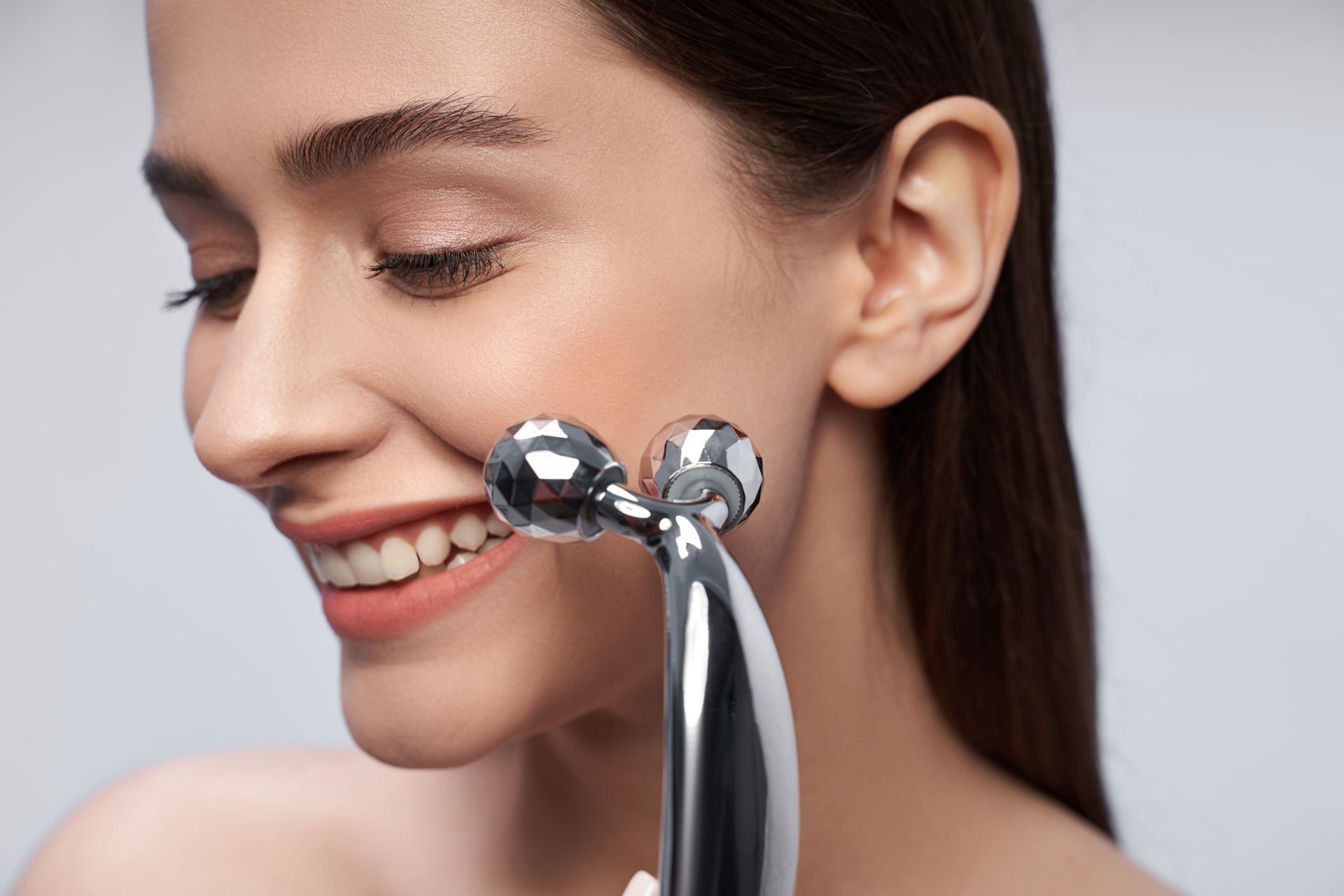 A woman is smiling while using a metal roller on her face.