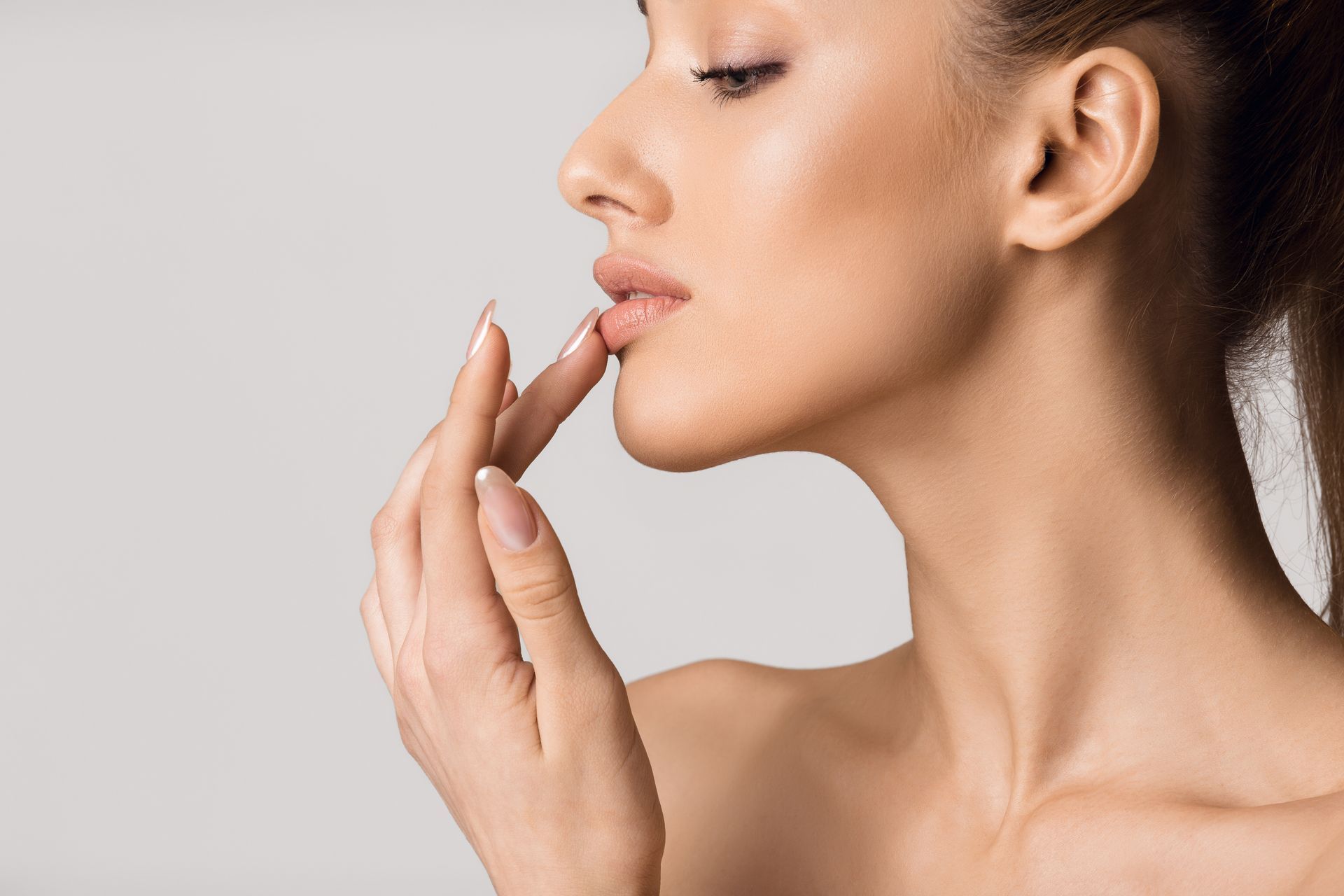 Woman with light skin touches her lips with her finger against a gray background.