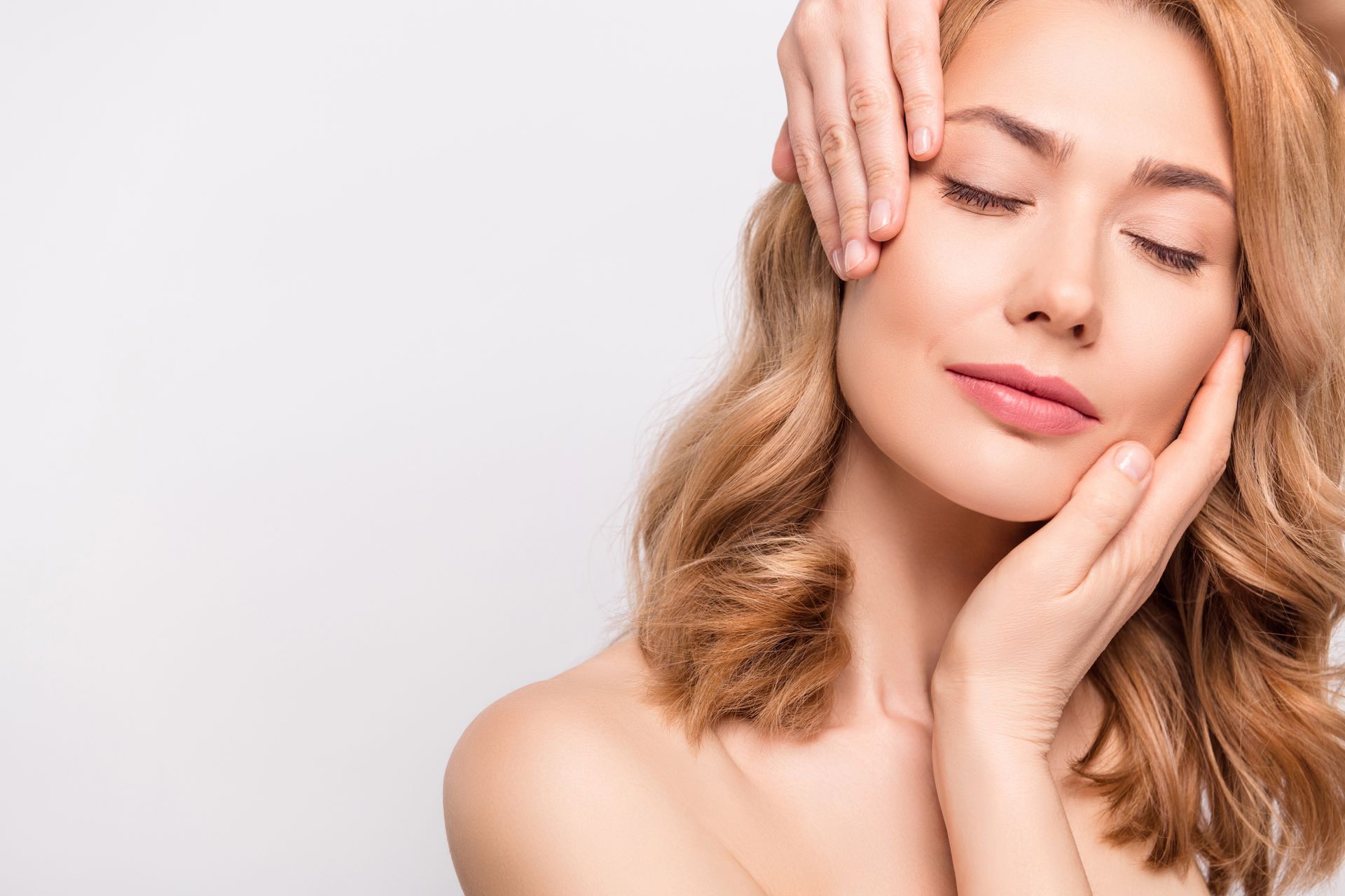Woman with closed eyes, soft skin, and hands gently touching her face; light background.