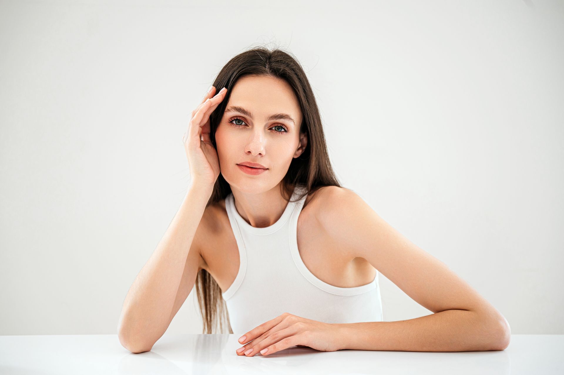 A woman in a white tank top is sitting at a table with her hand on her face.