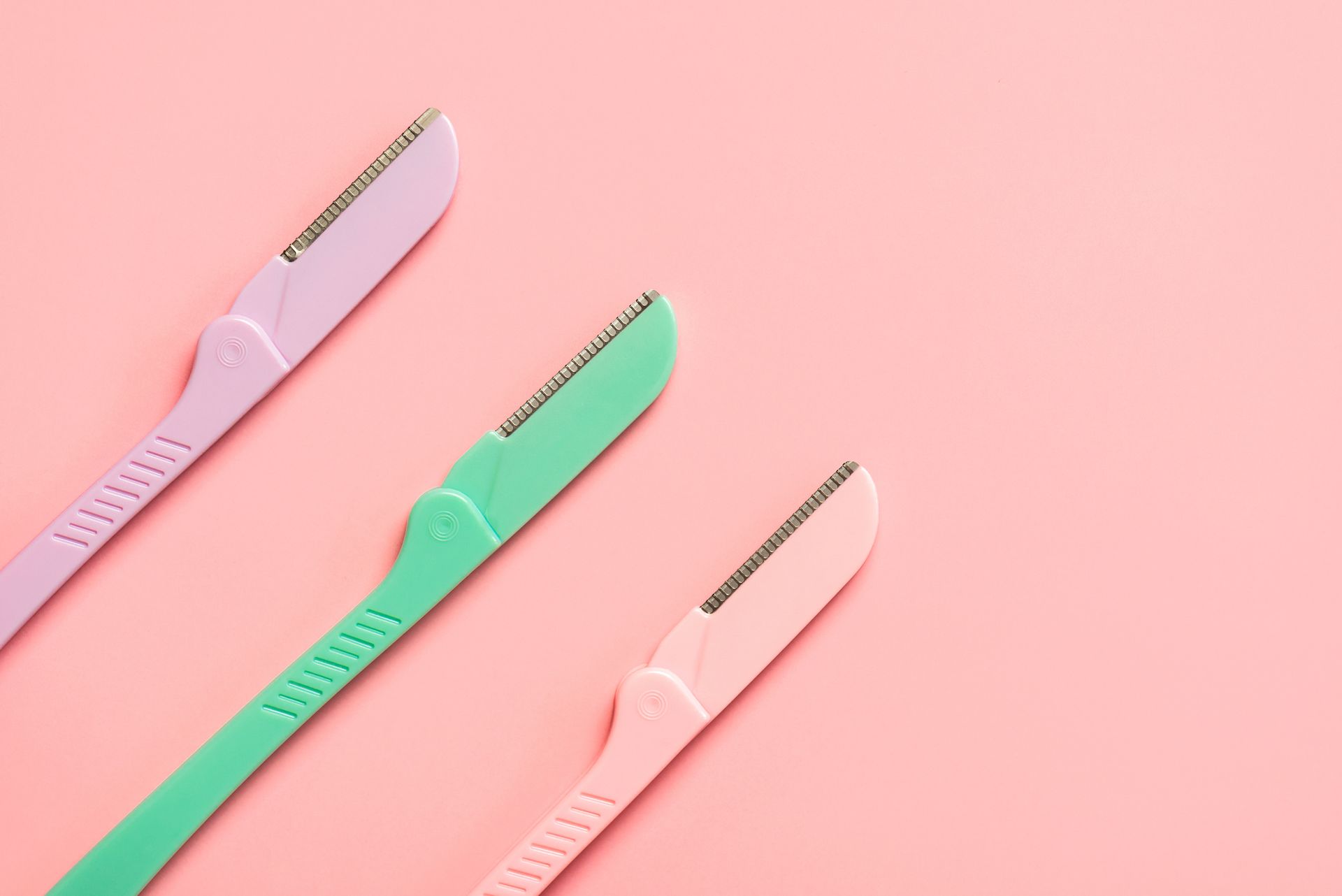 Three colorful eyebrow razors on a pink background; purple, green, and pink.
