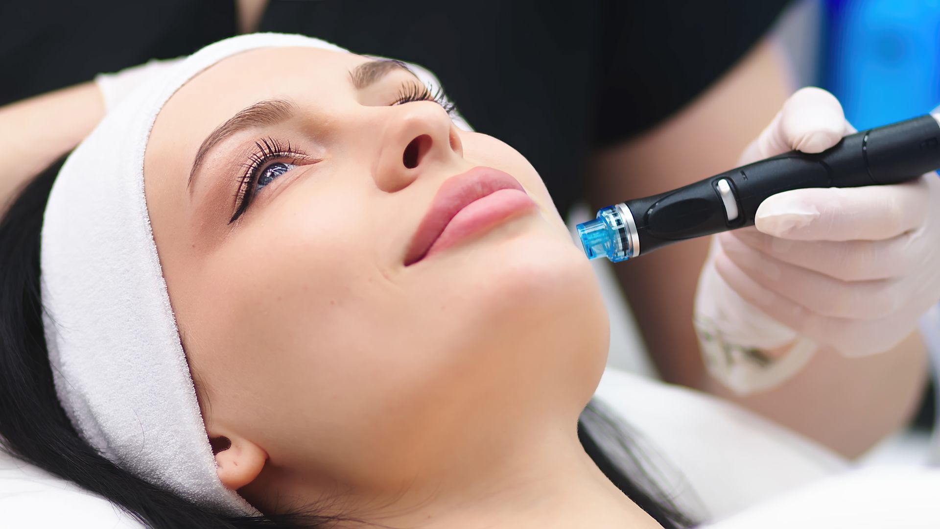Woman receiving facial treatment with a handheld device; white headband, gloved hand.
