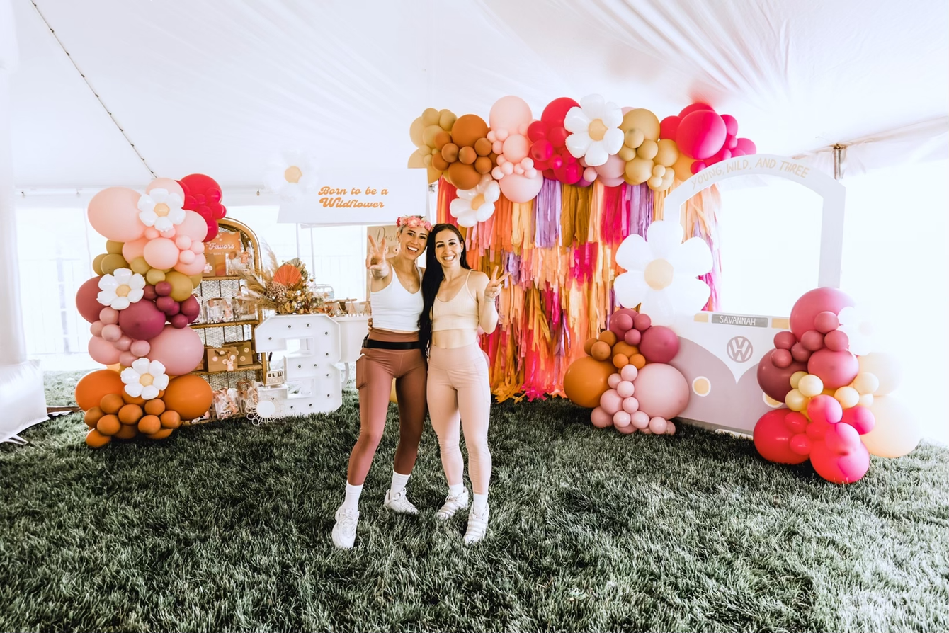 Two women are standing next to each other in front of a wall of balloons.