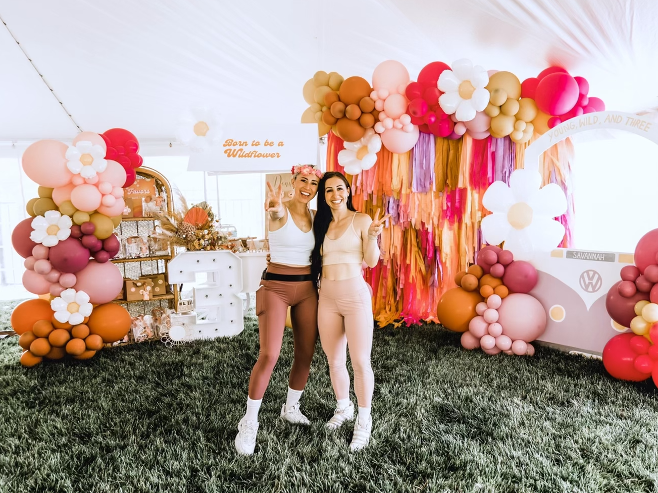 Two women in workout clothes pose in front of a colorful party backdrop.