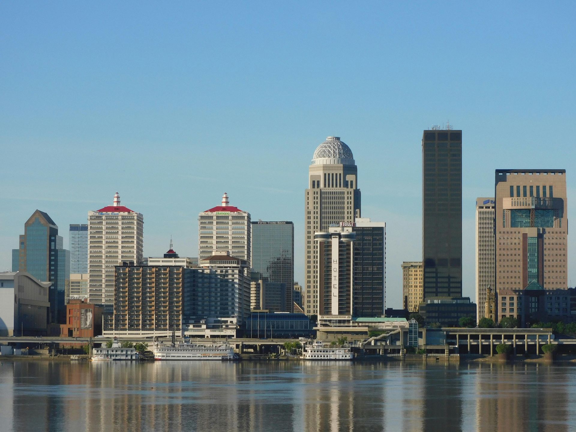 A scenic view of the Louisville skyline featuring the iconic PNC Tower dome, situated along the Ohio River under blue sky.