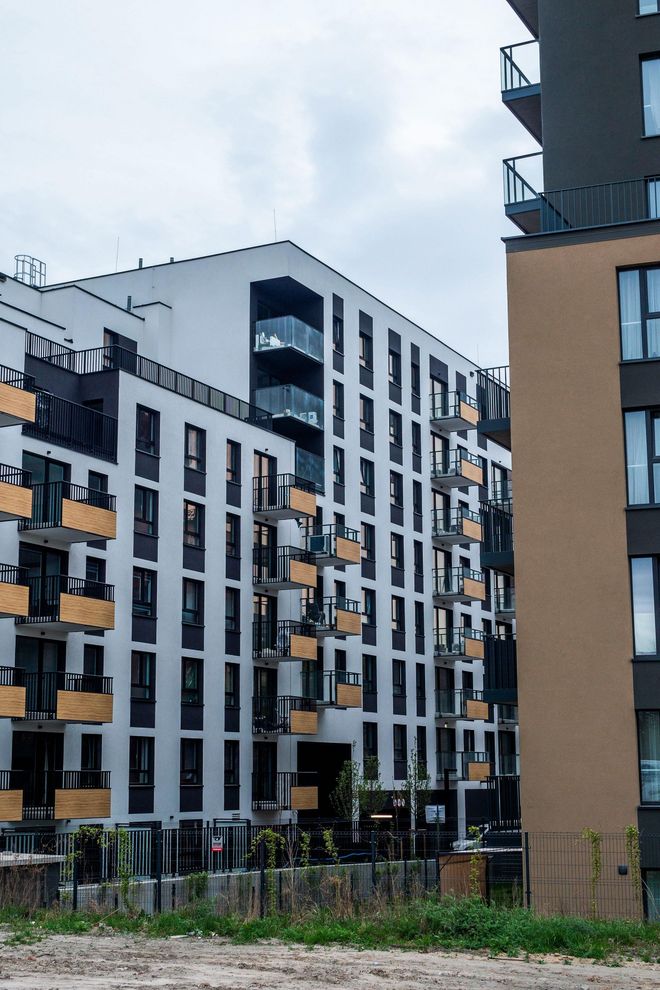 Modern apartment buildings with white and tan facades, featuring balconies and dark window frames under a cloudy sky.