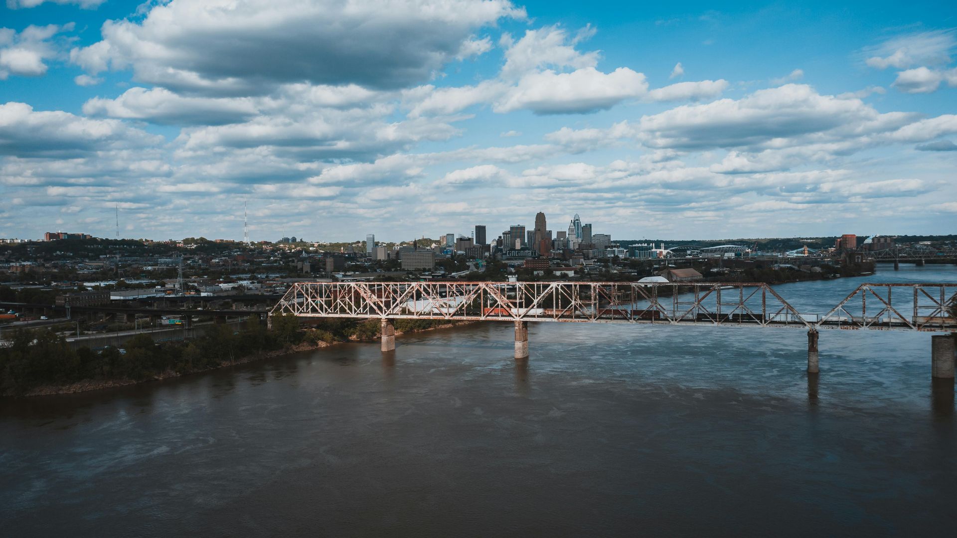 A wide-angle aerial view of a metal truss bridge spanning a wide river with a city skyline under a blue, cloudy sky.
