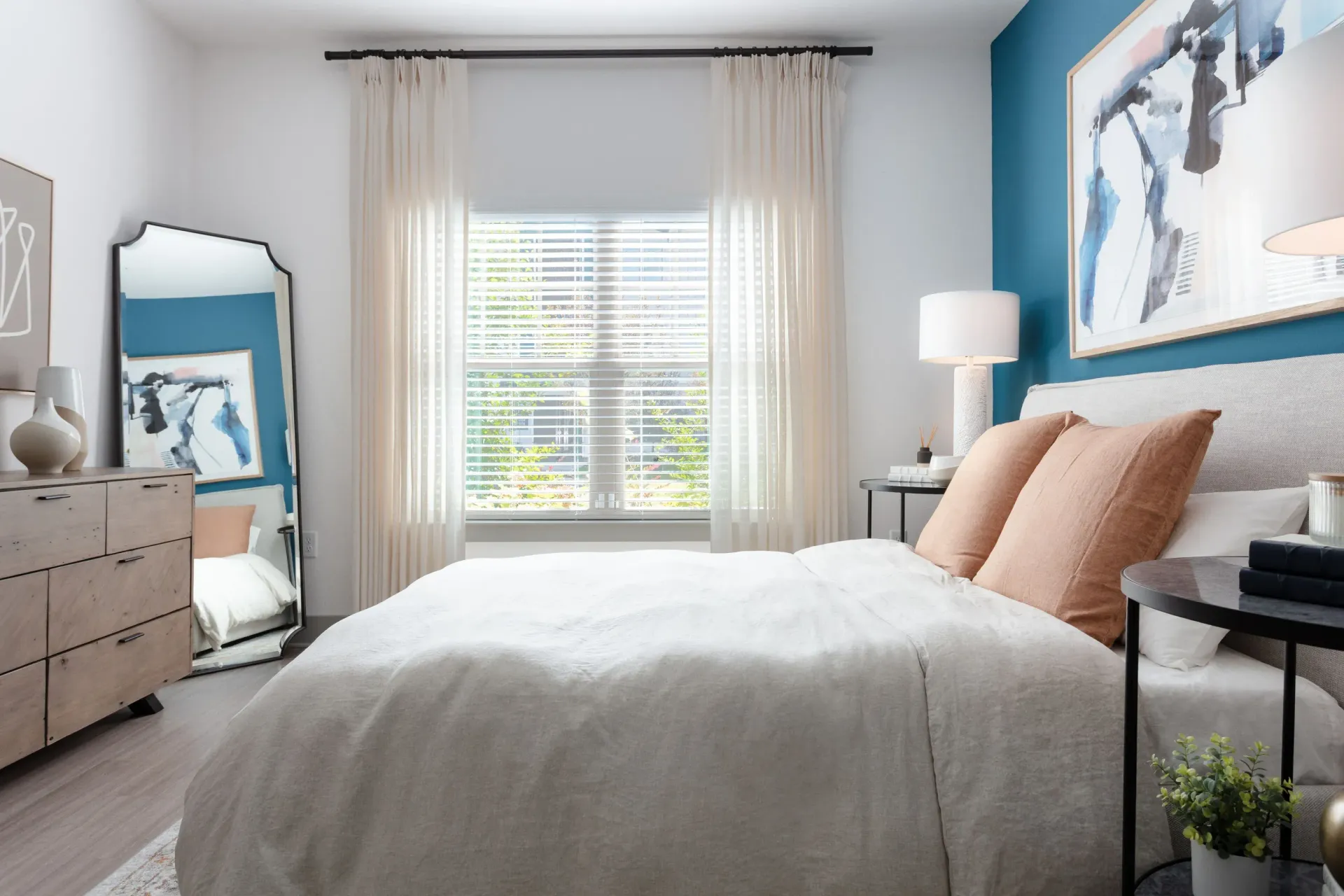 Bedroom with bed, peach pillows, dresser, and large window with sheer curtains.