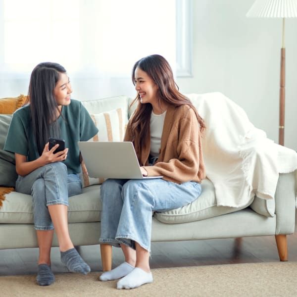 Two people sit on a light-colored couch, smiling while one uses a laptop and the other holds a smartphone.