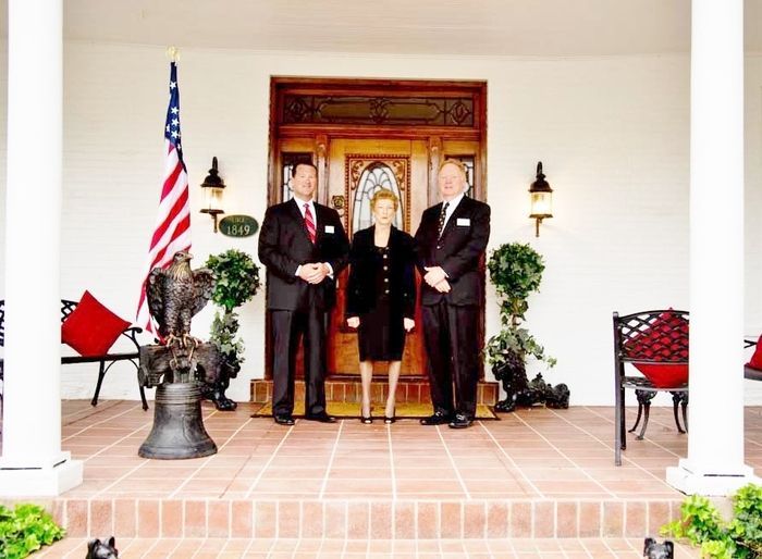 Three people in suits stand on a porch with American flag and ornate doorway.