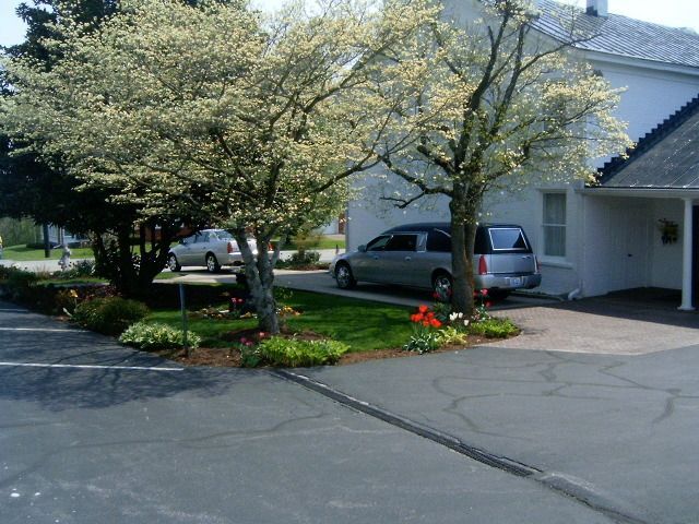 A hearse and a car parked in front of a white building with trees and landscaping.