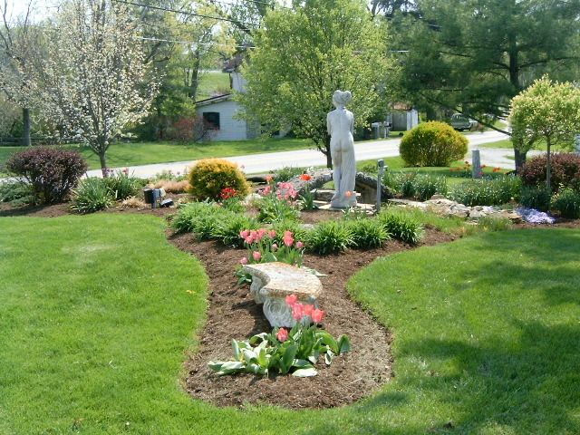 A well-manicured garden bed with a stone statue and colorful flowers, surrounded by green grass.