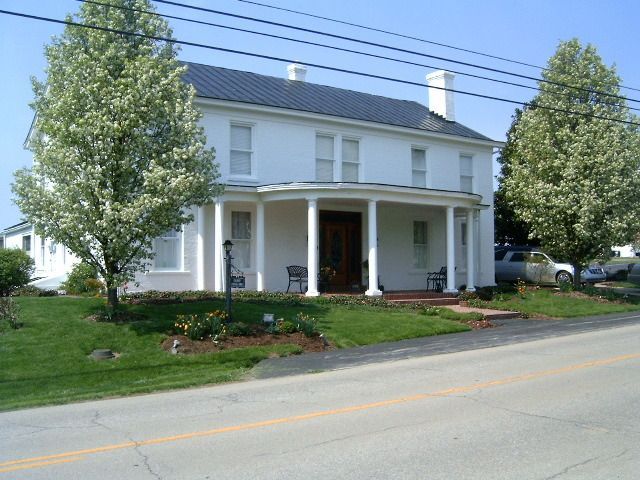 White, two-story house with porch and black roof, in front of green lawn and trees beside a road.