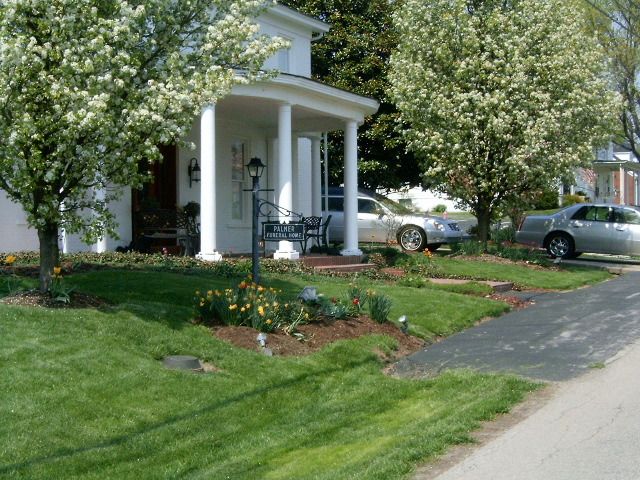 A white house with a porch, flanked by flowering trees and cars parked on the street.