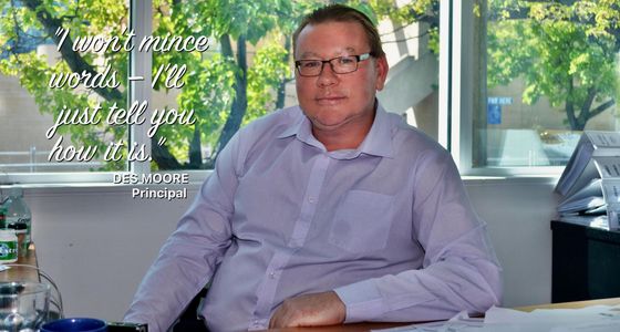 Man in glasses and a light purple shirt, sitting at a desk in an office.
