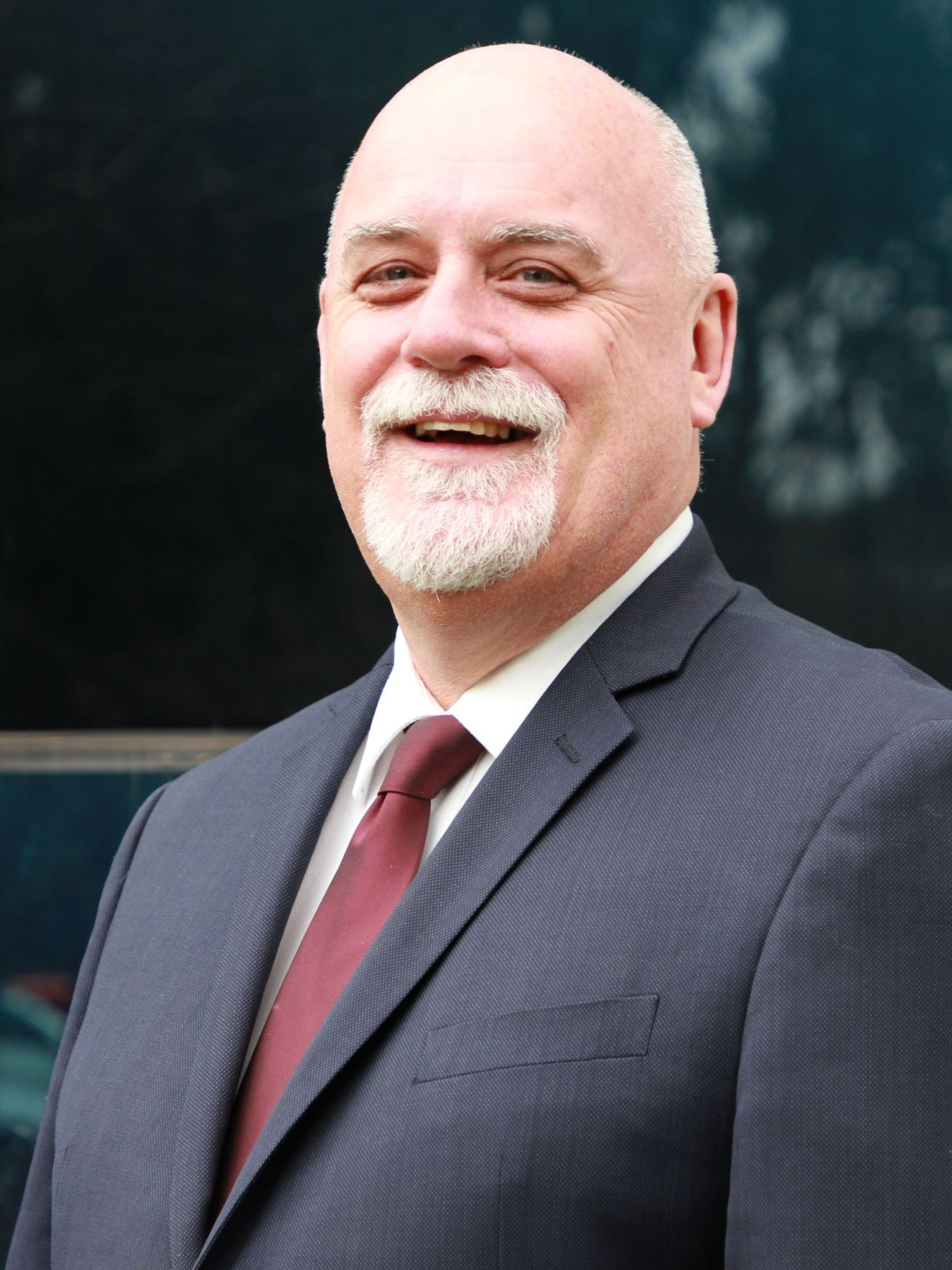 Bald man with a white beard smiles, wearing a navy suit and burgundy tie, outdoors in front of a dark building.