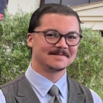 Man with glasses and mustache wearing a vest and tie; set against greenery.