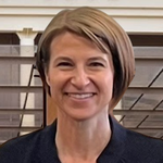 Woman with short brown hair smiles, wearing a dark blazer. Background of vertical blinds.