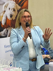 Woman speaking into a microphone, wearing a light blue suit. A dog poster is in the background.