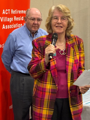 Woman in pink plaid jacket speaks into a microphone, man in blue shirt stands behind her. Red backdrop.