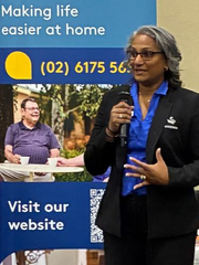 Woman speaking into a microphone, standing in front of a banner promoting home care services.