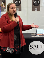 Woman speaking into a microphone, gesturing with one hand. Red jacket, dark patterned dress, 
