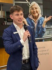 Man speaking into a microphone. Poster of older woman behind him. Indoor setting.