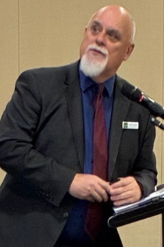 Man in suit with white beard speaking at a podium, holding pen and papers.
