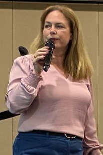 Woman in pink blouse holds microphone, speaking, indoors.