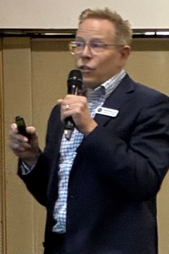 Man in blue blazer and glasses speaks into a microphone, holding a small object.