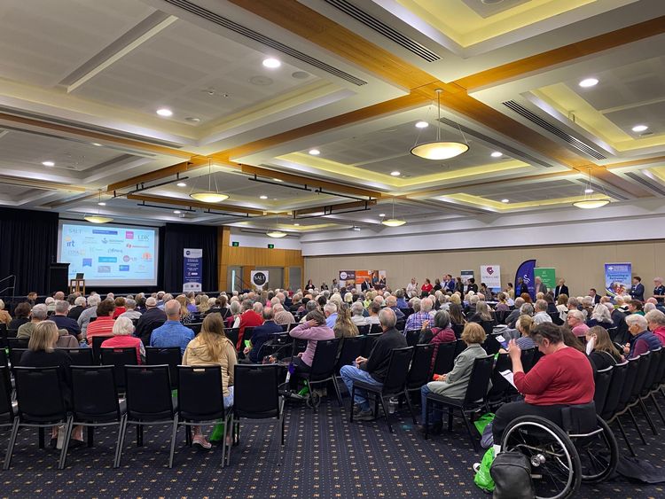 Large conference hall filled with seated attendees. A speaker on stage presents with a screen behind them.