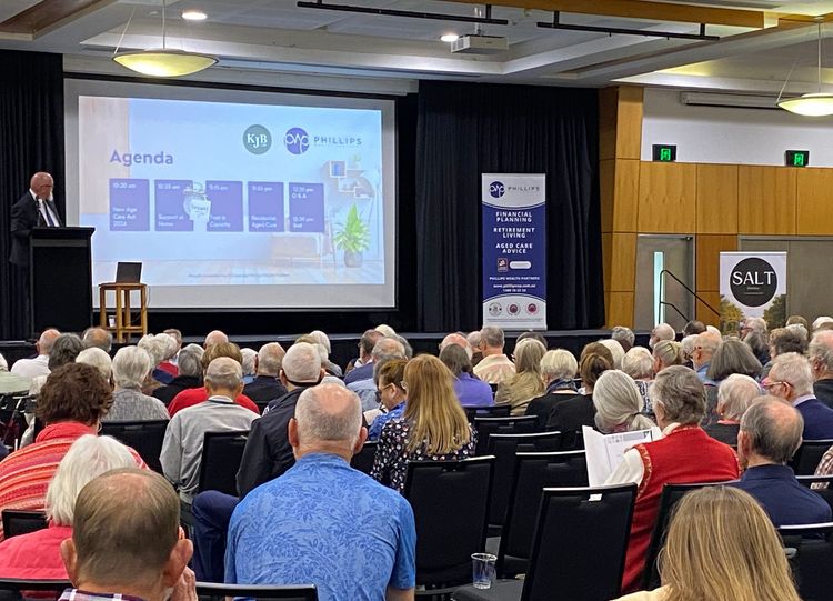 A man presents an agenda on a screen to a large audience in a conference hall.